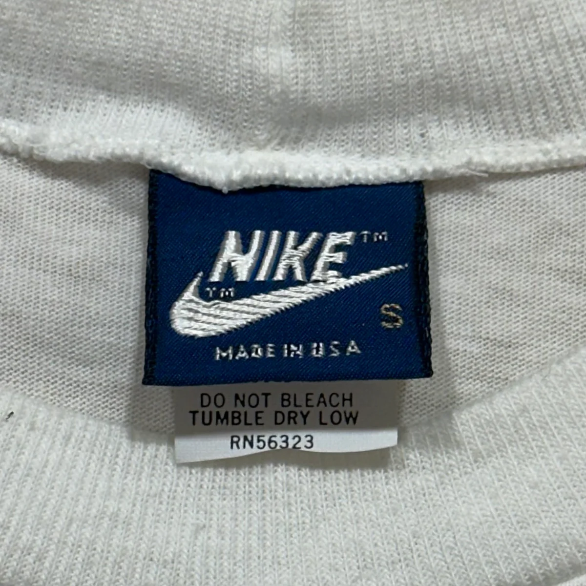 Vintage Nike blue neck tag with white Nike lettering and swoosh, MADE IN USA, size S, with the care label below showing RN56323
