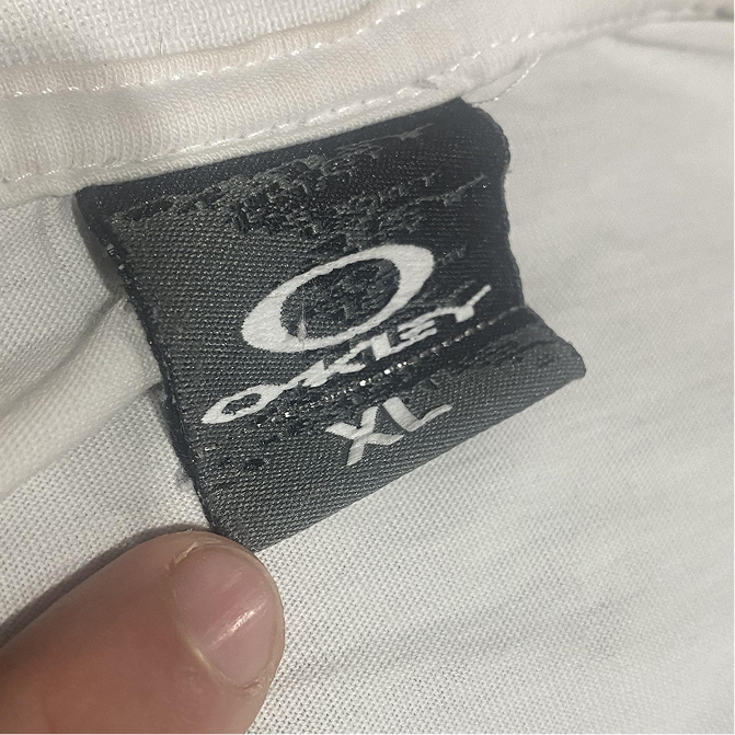 Black Oakley tag with metallic silver O ellipse logo and Oakley script, size XL, on a white shirt
